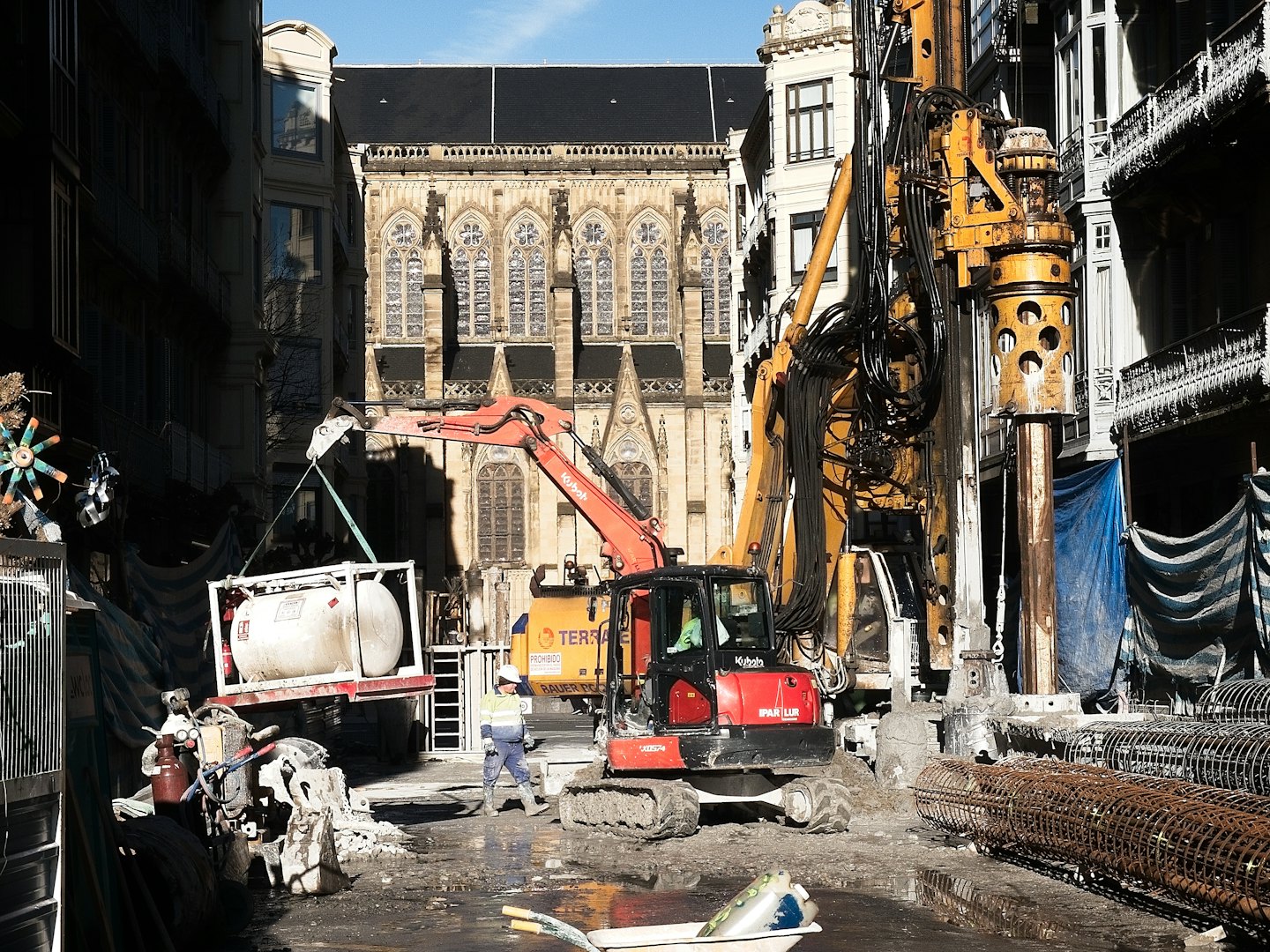 Construction worker supervises heavy machinery on a muddy urban site.